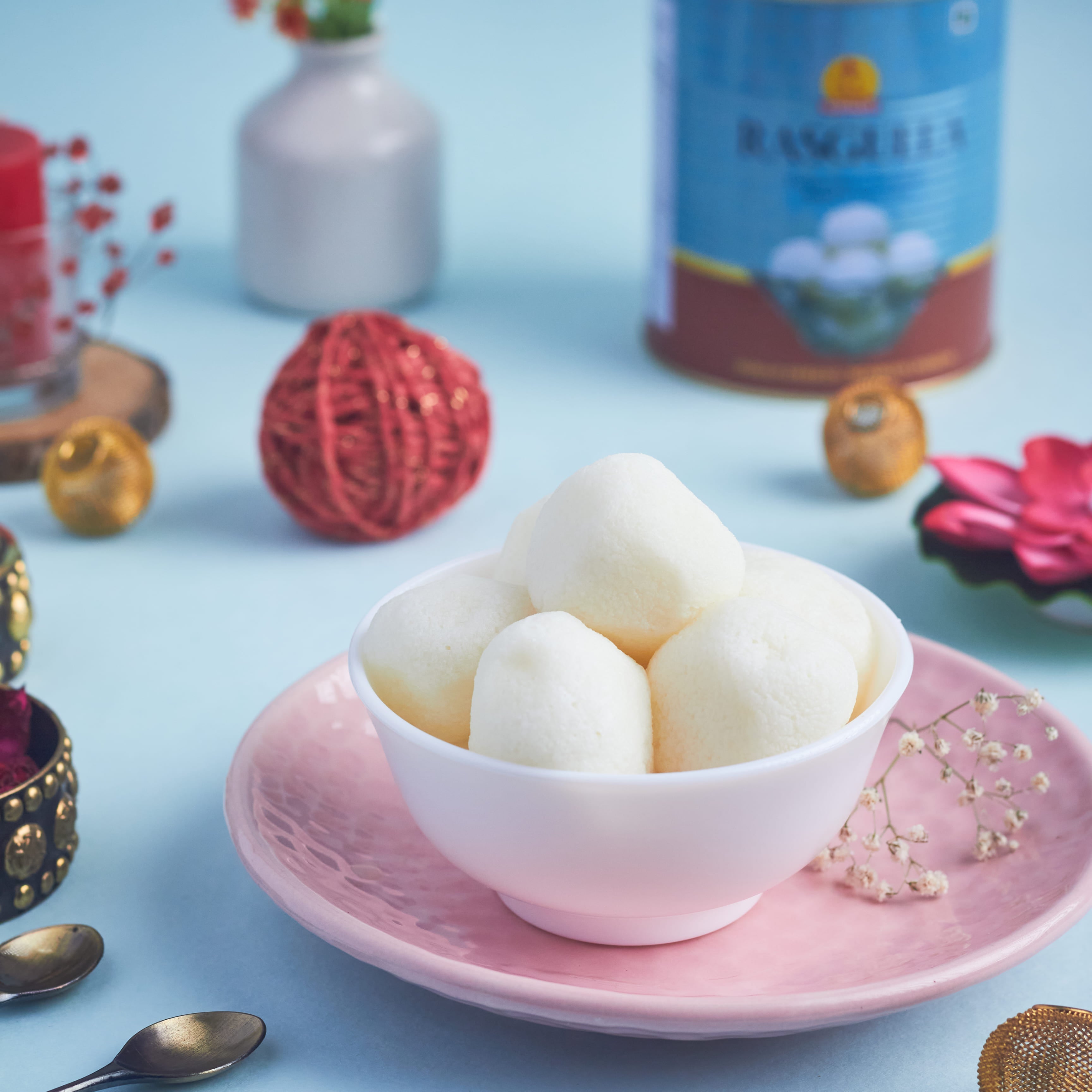 A bowl of soft spongy rasgullas styled on a light blue background with festive props and Rasgulla Tin in the background