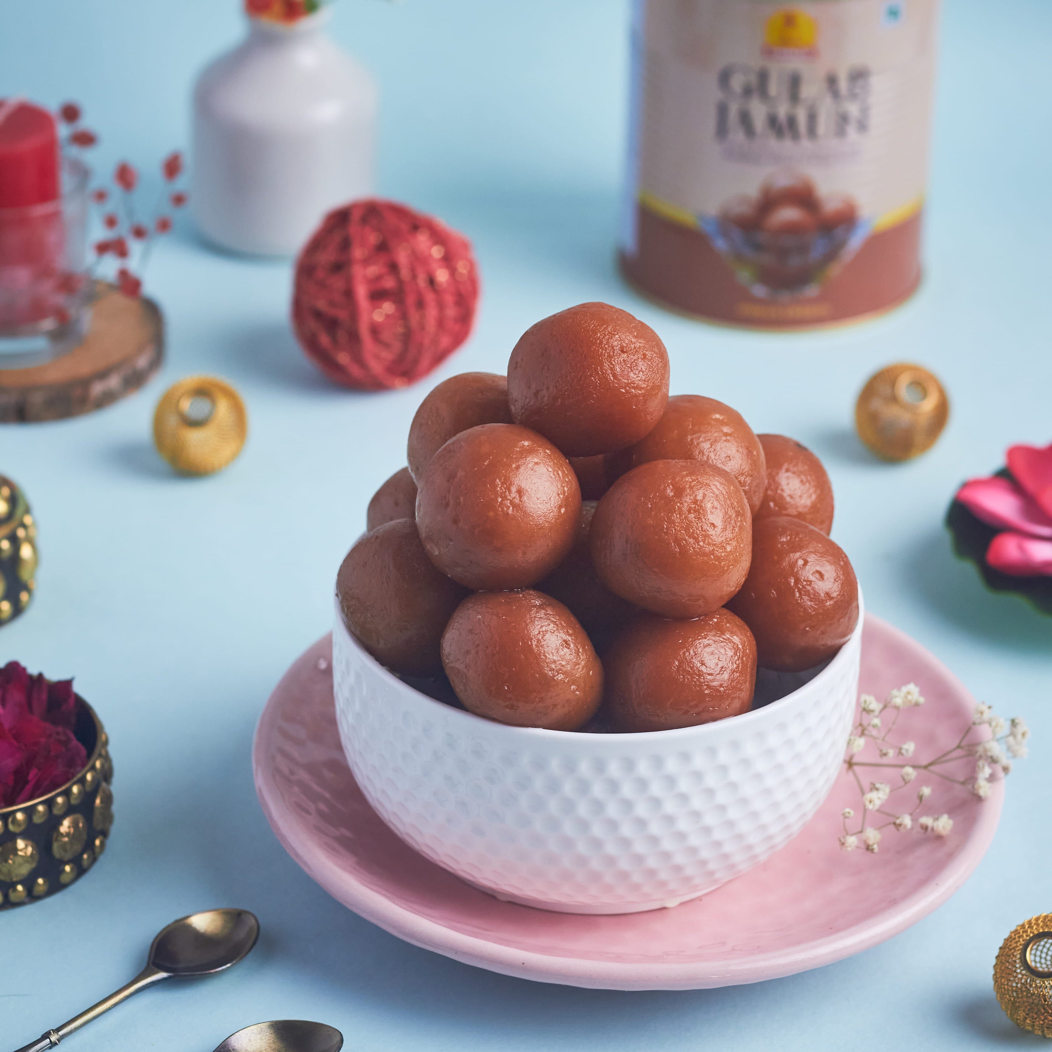 A bowl of gulabjaamun sweets on a pink plate, styled on a light blue surface with a can of Gulab Jamun, decorative props, and small spoons.